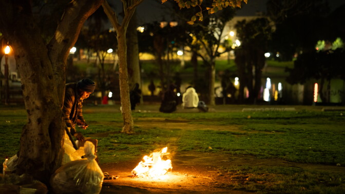 A man stands in front of a fire in a public park as other people sit on benches in the background.