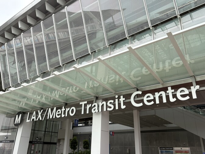 A black and white sign with an "M" representing L.A. Metro's logo followed by "LAX/Metro Transit Center" The sign reflects on the glass above it. 