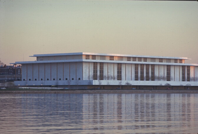 A large, low white building sits on the bank of a river. It is pictured from across the river.