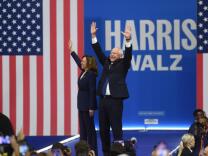 Two people both wearing suits wave to a crowd with an American flag in the background. 