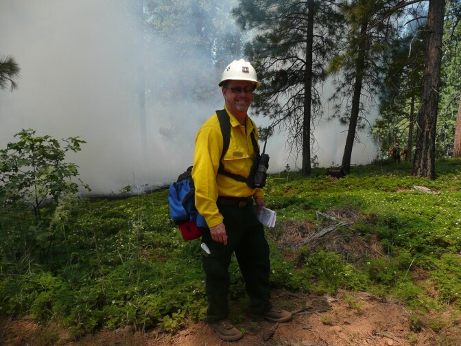 A man in a yellow safety shirt and white hard hat stands near a smoky area.