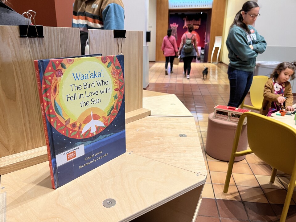 A picture book with the title Waa'aka': The Bird Who Fell in Love with the Sun. The title illustration is of a yellow sun with silhouette of a white bird. There is a family sitting at a table with yellow chairs in the background. 