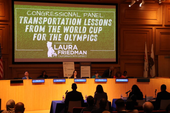 A group of five people sit at a dais. Behind them is a projection that reads: "Congressional Panel: Transportation Lessons From the World Cup for the Olympics." The bottom of the projection has the text: "Congresswoman Laura Friedman, California's 30th District."