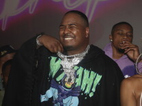 Drakeo The Ruler is smiling widely, holding up a pile of thick platinum chains that hand around his neck. Other people mill about next to him and behind him. 