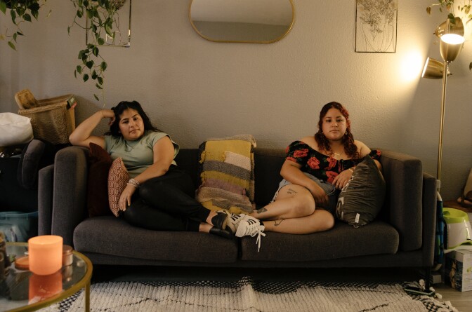 Two young women lounge on opposite sides of a sofa in a dimly lit room with a lamp on the right side. A mirror is above the sofa.