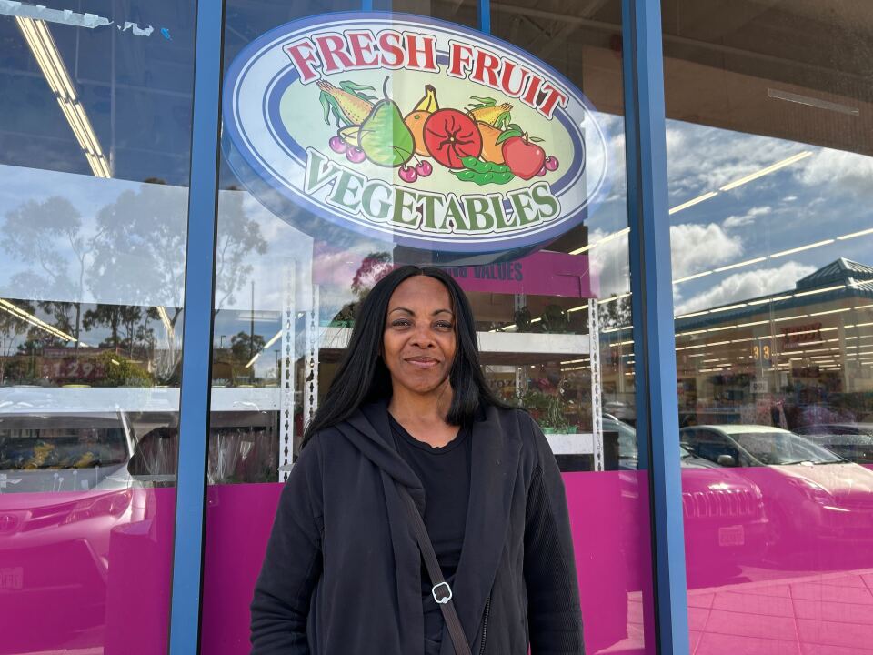 A woman with long black hair standing in front of a store front with a sign that says "Fresh Fruit Vegetables"