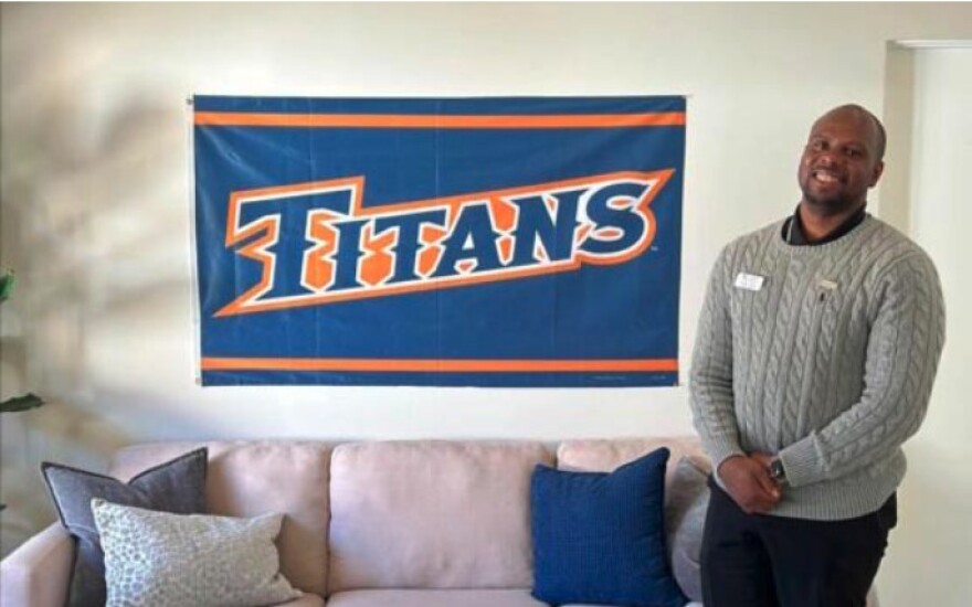 A man who appears to be Black and in his 30s stands smiling next to a blue and orange banner that reads "Titans." He is in a living room next to a couch. He wears black pants, and a grey sweater over a black collared shirt.