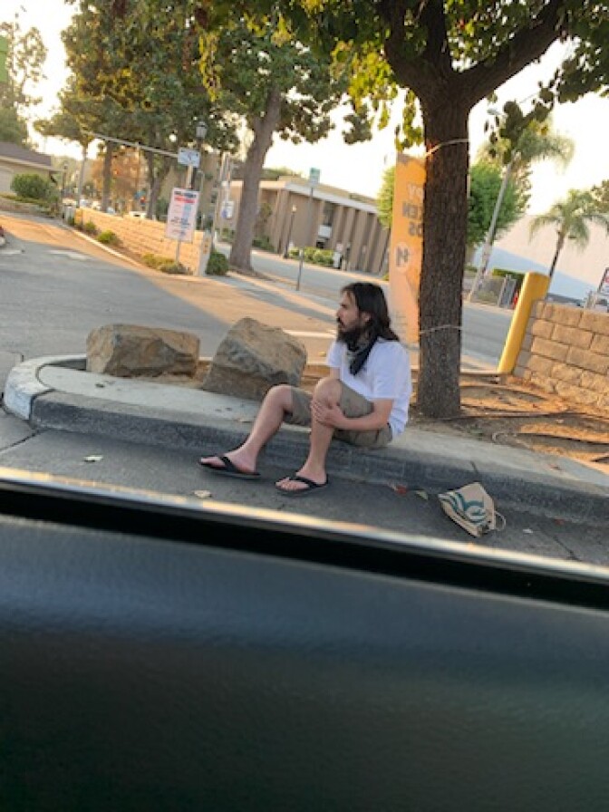 Frank Campos sits on a curb beneath a tree. He wears shorts and a white t-shirt and flip-flops