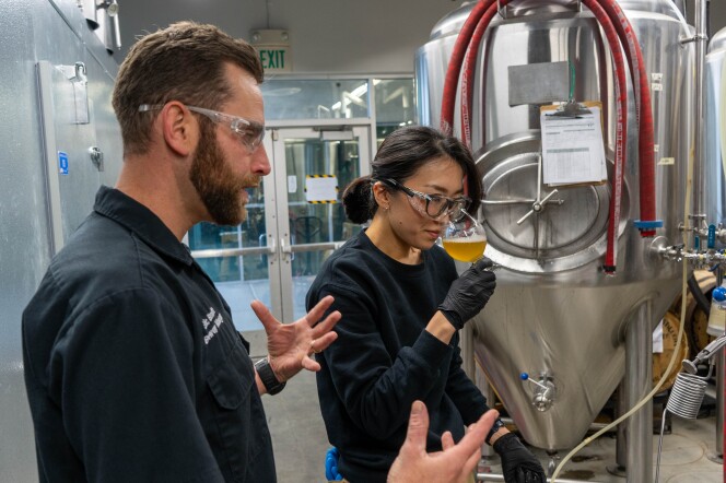 A male presenting person and a female presenting person wear dark blue clothes. They stand next to metal vessels. The female presenting person holds a glass with bubbly, golden liquid.