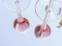Two glasses of rosé wine are shown reflected on a glass table. 