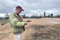 A middle aged man with light skin wears a light green jacket and red baseball cap and gestures toward the ground while standing on a dirt lot on a rainy day. 