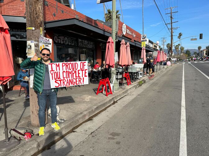 A man in a down coat and sunglasses holding a sign that says "I'm Proud Of You Complete Stranger!" and standing on the sidewalk on the course of the L.A. Marathon