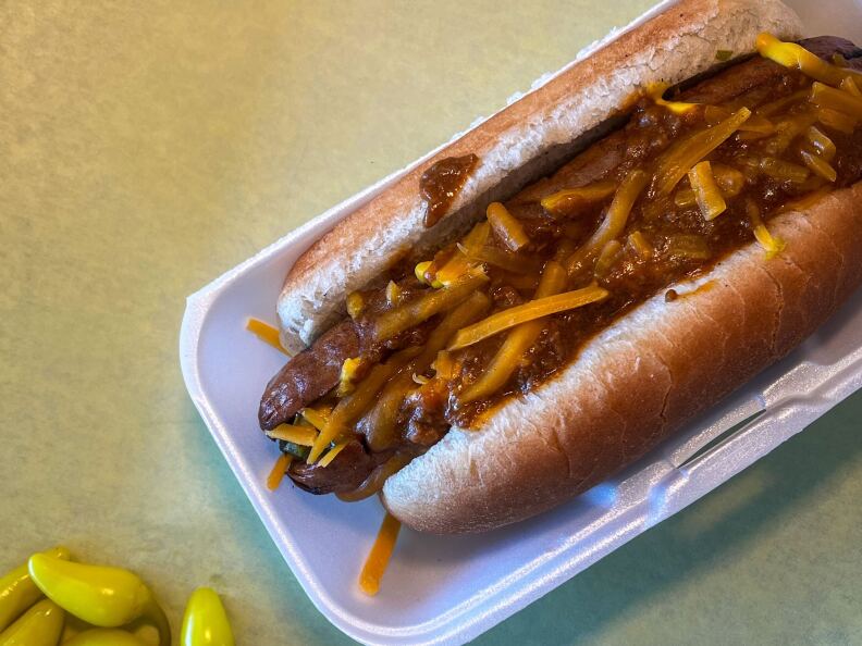 A hot dog with a light brown bun sits on a rectangular white styrofoam plate. The hot dog itself is sliced practically down the middle and is filled with orange cheese then topped with brown substance then topped with more cheese. In the bottom left corner is practically shown pile of little yellow chile peppers.