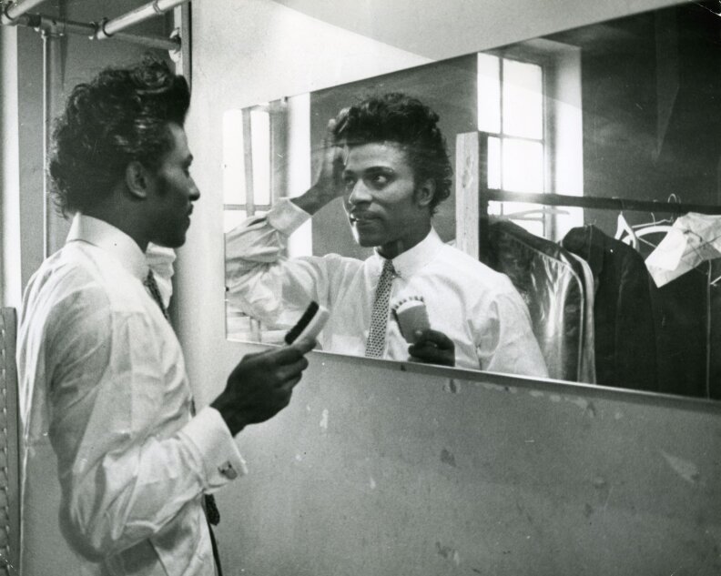 Little Richard adjust his hair in a mirror. He holds a brush and wears an untucked white button-up shirt, along with a tie. Other suits are seen on a clothing rack in the mirror's reflection.