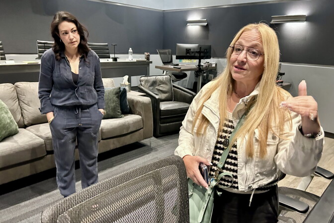 Two female presenting persons stand inside a room painted gray. Several couches are behind them.