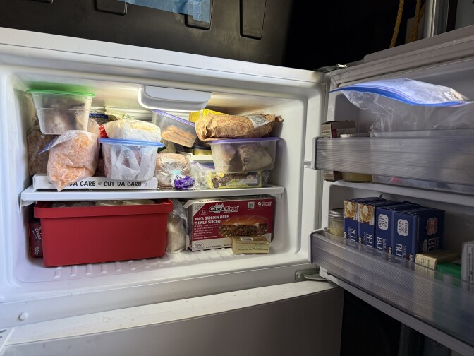 A freezer is stuffed with a variety of bags, bins and boxes. The door is lined up with a variety of butters. 