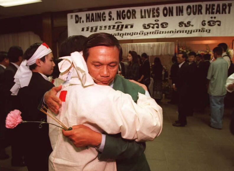 An Asian man is shown hugging someone. They are surrounded by other people, several men wearing suits. A banner hangs in the background reading "Dr. Haing S. Ngor Lives On In Our Hearts."