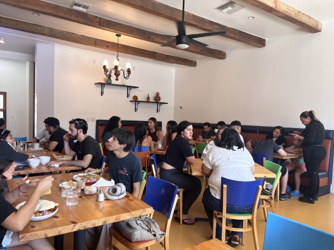 A full restaurant of people eating dishes at various tables. A server writes down in a notepad in the background as they talk with a table of guests.