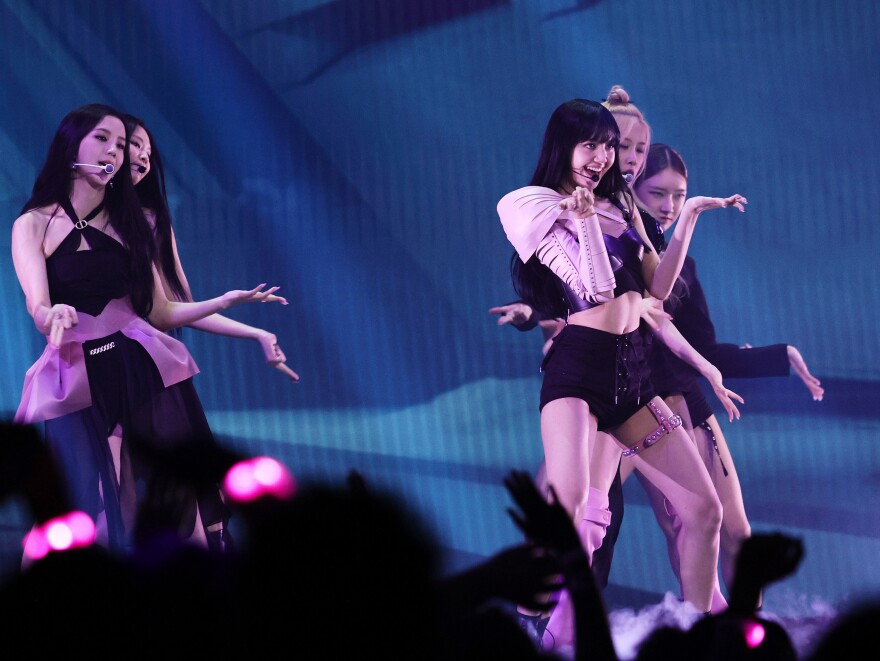 A group of Asian woman in black and pink dance outfits perform on stage with mics