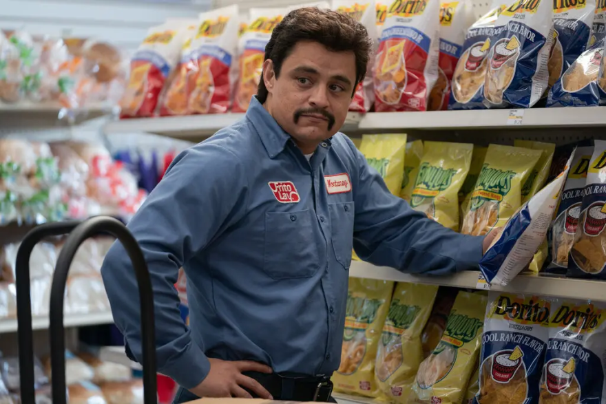 Jesse Garcia stands in the chips aisle of a grocery store. He is placing bags of tortilla chips on the shelves. He wears a blue jumpsuit with the emblem of Frito Lay on the right side of his chest.