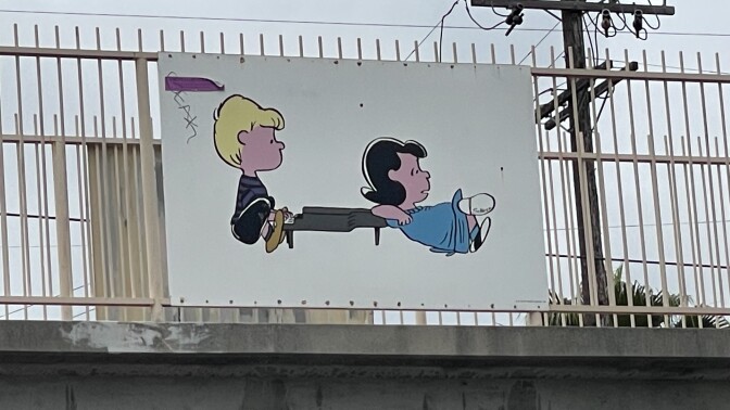 A white panel, featuring the Peanuts comics characters, is affixed to the tan railing of a bridge. The panel features cartoon drawings a young girl with black hair and blue dress reclining on a small piano, being played by a boy with blonde hair wearing a gray shirt, shorts, and tan shoes. 