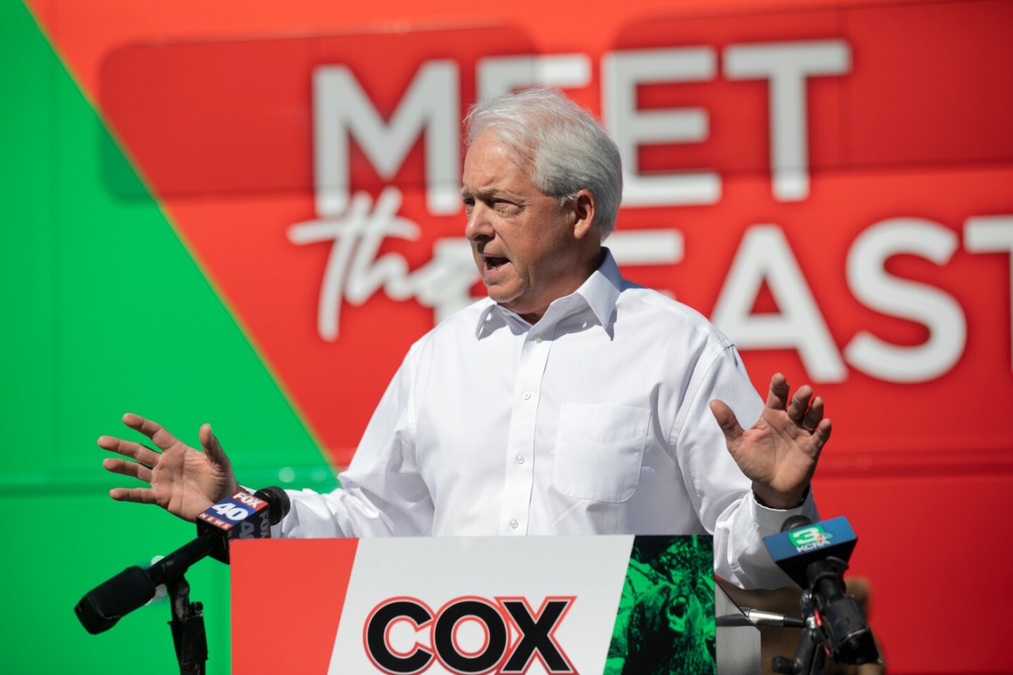 John Cox stand a a lectern with his hands raised to chest level as he speaks in from of a bright green and red backdrop. The sign on lectern reads: Cox