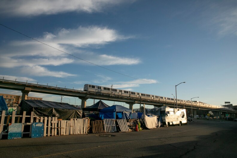 An elevated train passes over an encampment of tents and tarps and fences made of pallets.