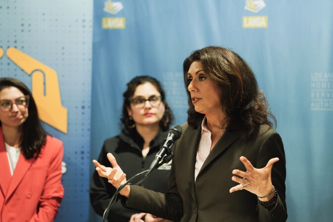 A woman speaks at a podium as two women look on from behind.