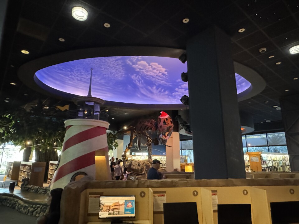 The interior of a creatively designed library or educational space with a strong thematic and immersive atmosphere. The ceiling features a large oval-shaped recessed section illuminated with a soft purple glow, depicting a textured cloud-like pattern that gives the impression of a sky scene. A row of computer stations is visible at the bottom of the image, housed in a structure resembling natural rock formations, adding to the thematic design.