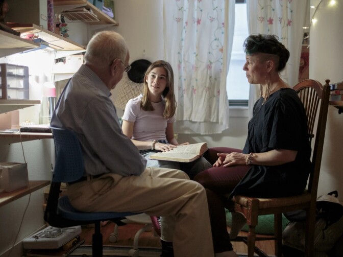 A young, white teenage girl sits with an older white man and white woman, with a Jewish prayer book open on her lap. The older woman has partially shaved hair, the test dyed blue. The girl is dressed in a pink shirt and jeans. The man wears khaki pants and a blue dress shirt. 