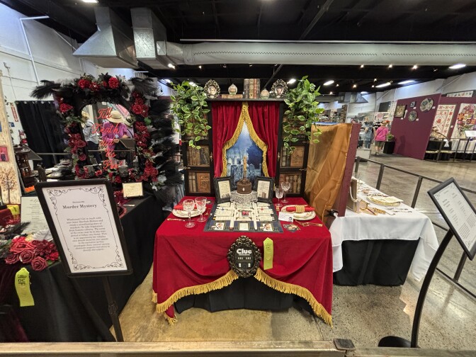 A view of a mini sized table with two table settings and a small background that features a faux window looking out onto a graveyard with red curtains with gold tassle trim.