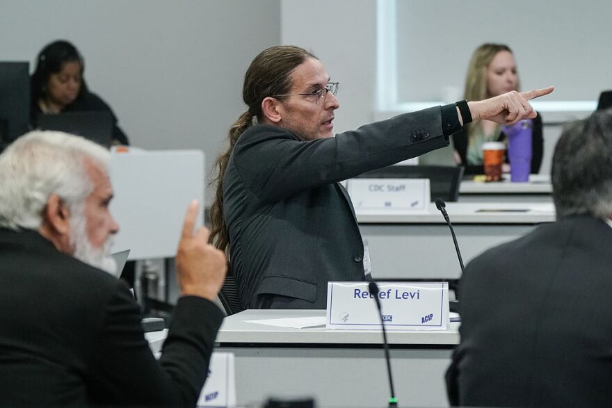 A man wearing a dark grey suit is pictured in profile. He is pointing his finger, arm outsretched, while speaking. There are two men and two women seated around him.