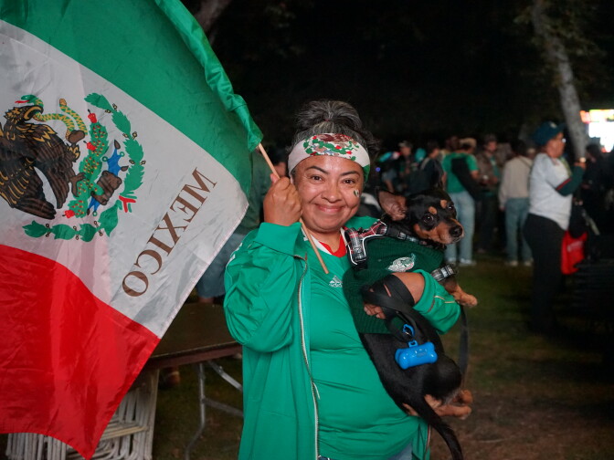 A medium skin toned woman with grey hair pulled back in a ponytail wears all green and clutches a small black dog. She holds a large Mexico flag and wears a bandana with the Mexican flag's pattern around her head.