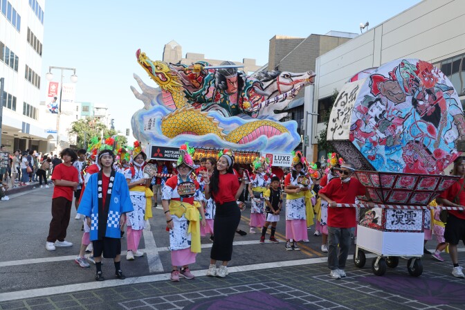 People in red shirts and others in Japanese robes walk alongside two parade floats.