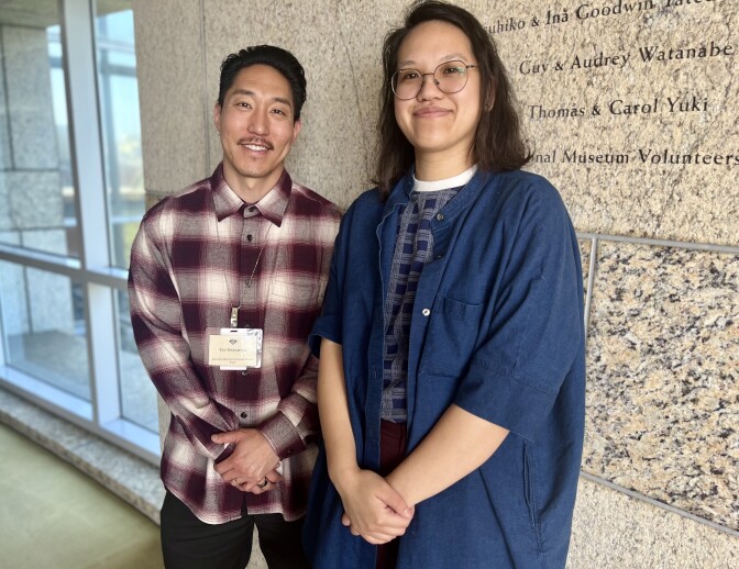 Two people, one in a maroon plaid shirt and another in a blue shirt, stand against a granite wall for a portrait. 