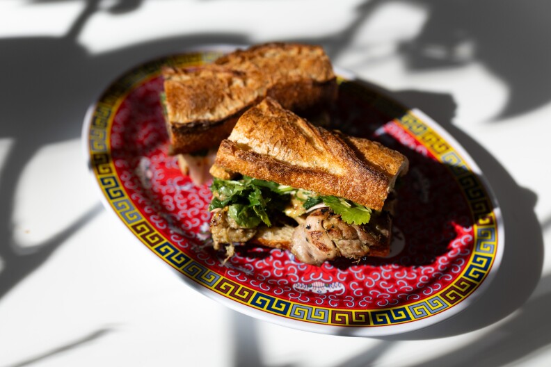 A photograph of a sandwich on an oval shaped plate embelished with traditional Asian designs: The sandwich has been made on a light brown baguette, and peeking out of the side are slivers of grilled pork topped with leafy greens. The plate sits on a white surface, and various backlit shadows overlay the plate of food.