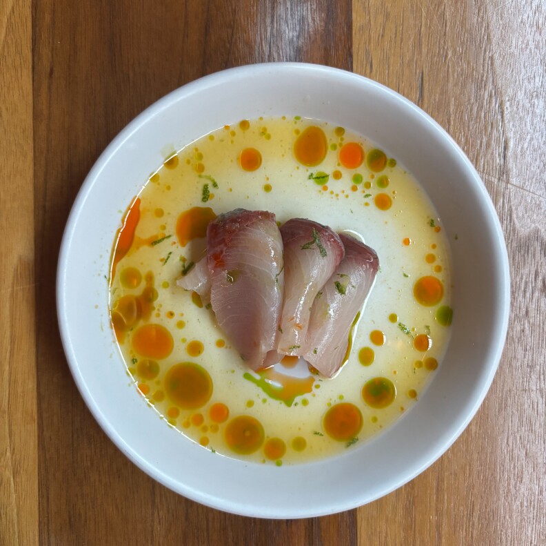 A shallow white bowl containing a delicate arrangement of raw fish slices resting in a citrusy broth dotted with vibrant green and orange oil droplets, all set on a wooden table