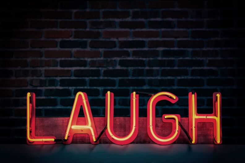 A neon sign says, 'LAUGH' as it stands in front of a brick wall.