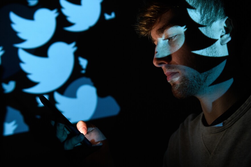 In this photo illustration, the blue bird logo for the Twitter social media network is projected onto a man in a dark room on Aug. 9, 2017 in London, England. Multiple images of the blue bird logo appear on a dark background behind him. He holds his phone, illuminating his face.