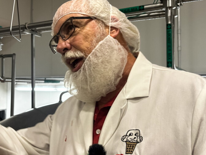 A man with a long white beard, wearing glasses, a hairnet, and beard net, gestures while speaking, wearing a white lab coat embroidered with the Thrifty Ice Cream logo.