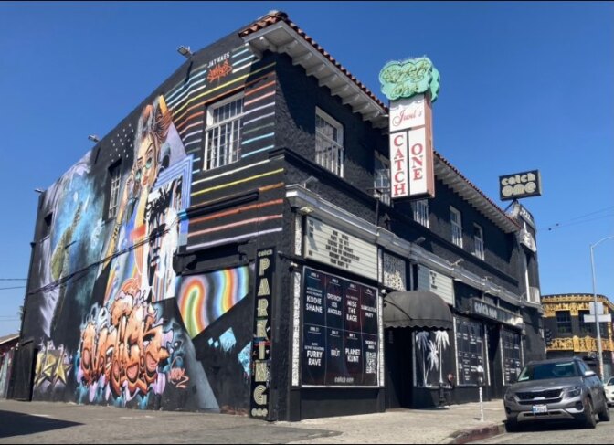 A large multi-colored building sits on a street corner; There are rainbow colored murals on one side; a sign above the entrance says Jewel's Catch One.