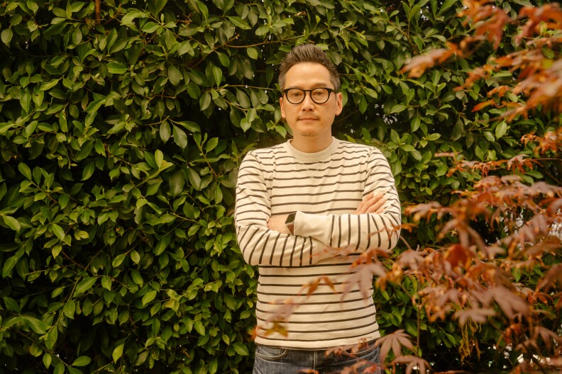 Glenn Kaino stands, arms folded, in a white and navy striped shirt, with a plant with green leaves behind him, one with red leaves in the foreground.