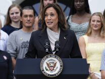 A female-presenting person with brown hair and medium-dark skin tone wearing a navy blazer and white blouse speaking into a microphone at a podium which bears the presidential seal. 
