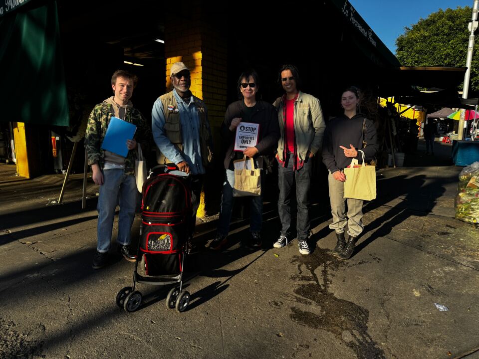 A man in a camouflage sweatshirt and blue jeans stands next to a man in a cargo vest with a stroller. He stands next to a man in black sunglasses and a black shirt who holds a sign that says "Stop Employees Only". He stands next to a man in sunglasses, wearing a green jacket, plaid shirt and red undershirt. A woman stands next to him in a black sweatshirt and holds a white tote bag. They pose for a picture together.
