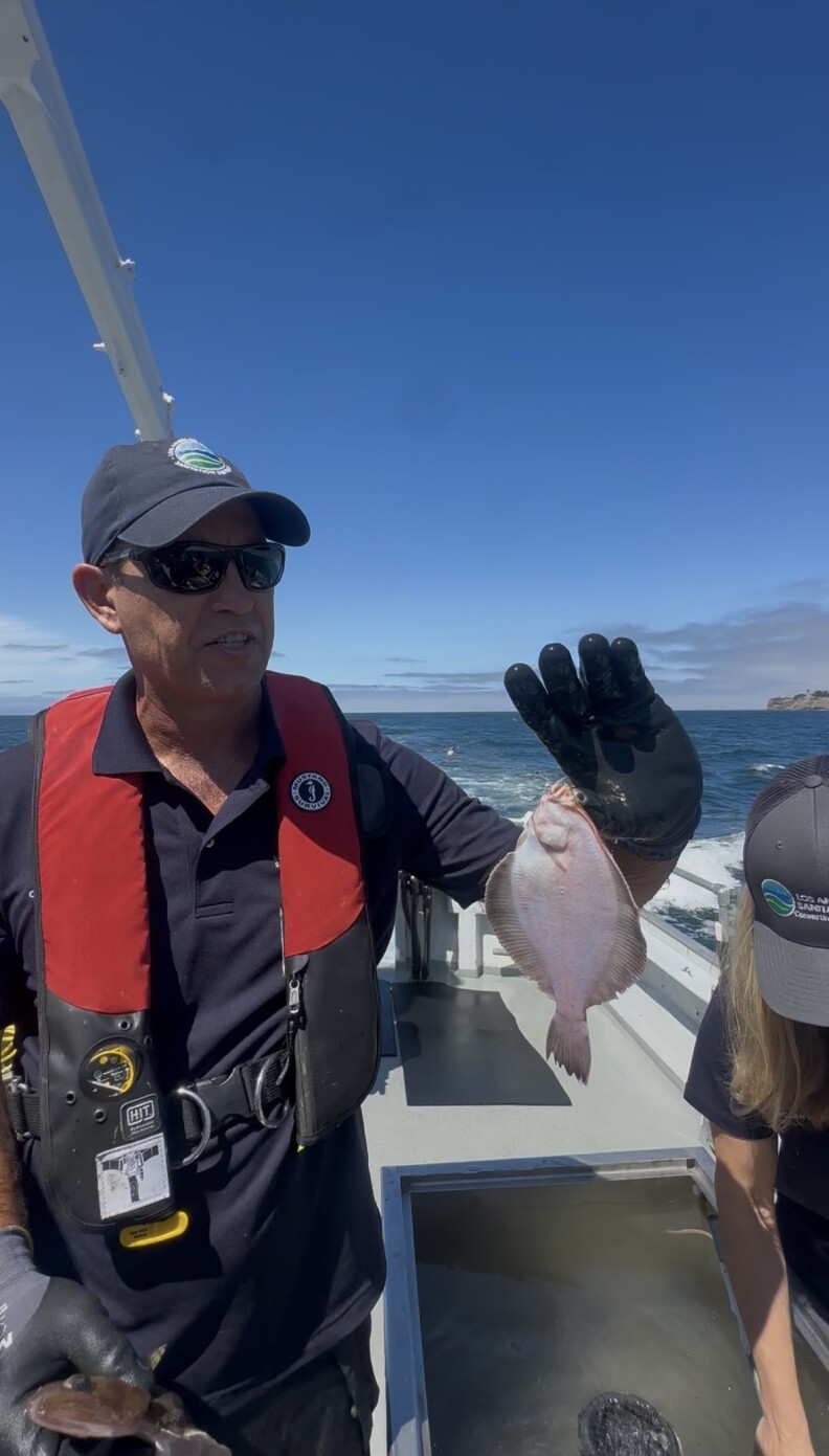 A man with medium skin tone and a life vest hooks a fish on his thumb.