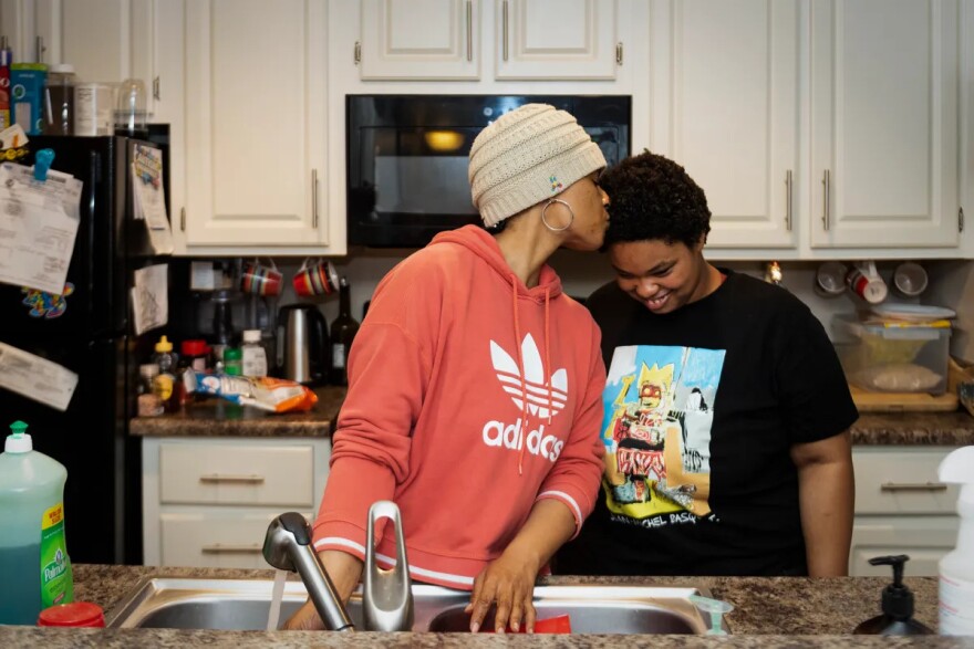 A Black woman and her teenage daughter stand at a kitchen sink in their home. The mother kisses her daughter on the top of her head as the girl smiles.