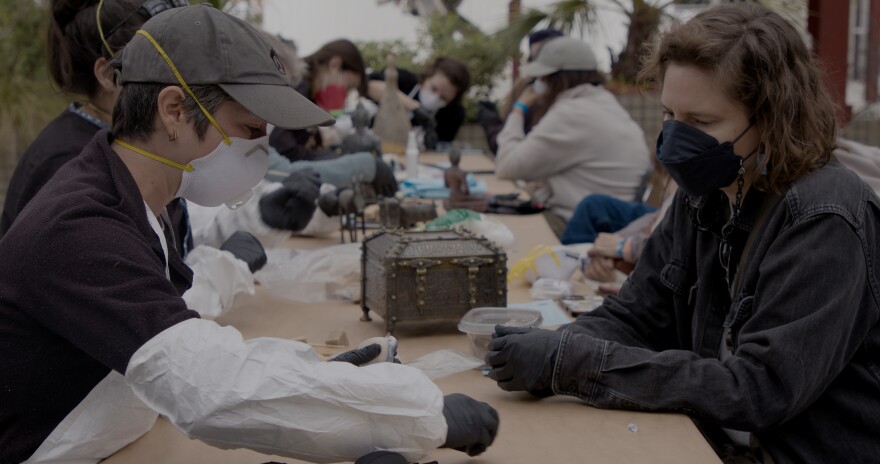 A conservator consults with a community member about their damaged object, assesses the condition, and proposes potential next steps. Both wear masks and sit across from each other at a table.