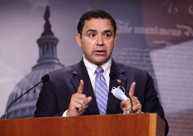 A man who appears to be Latino and in his 50s or 60s stands behind a podium speaking. His expression is serious. He gestures with both hands. He is wearing a navy suit and blue tie. 