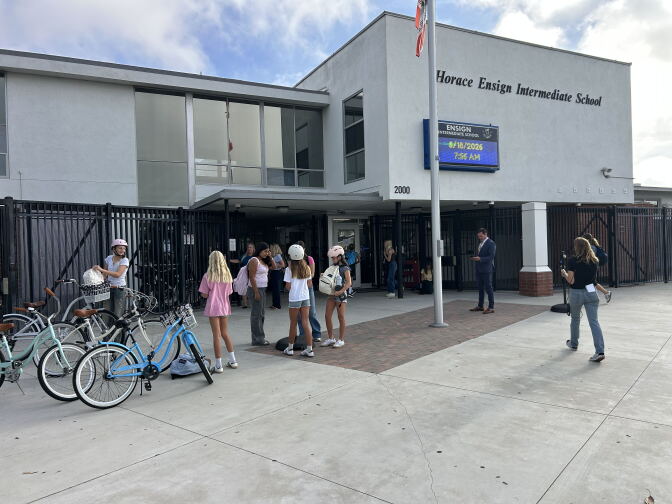 In front of a school building, a group of students stand outside. Three bicycles are parked out front. 