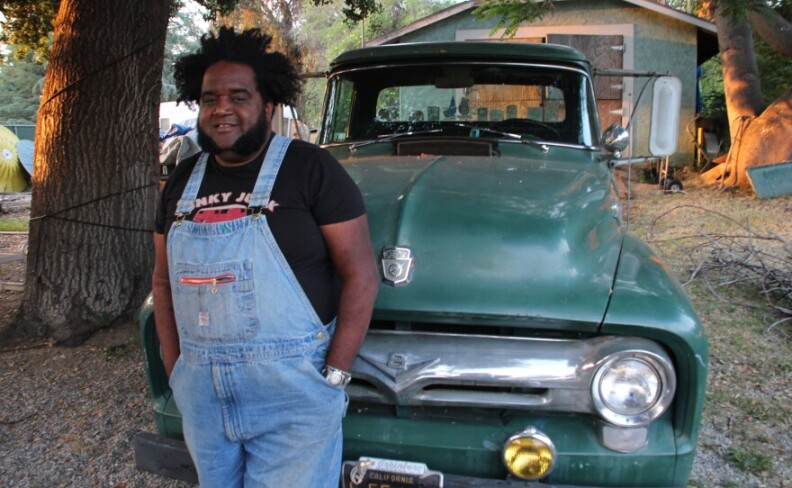 A man with dark skin tone wearing light blue overalls and a black T-shirt Jstands in front of a vintage green truck.